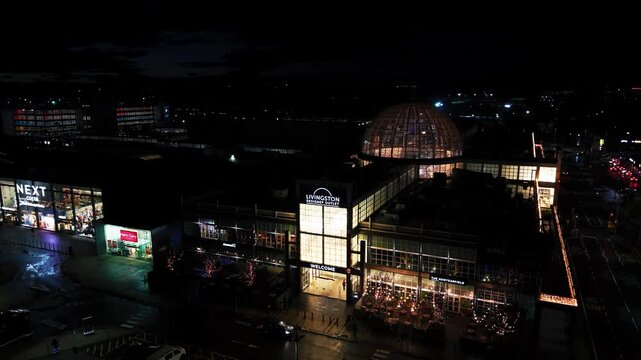 Aerial drone footage at night showing the main entrance of Livingston Designer Outlet with illuminated signage, retail stores, and surrounding urban area in Livingston, Scotland