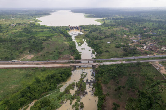 Aerial view of the Asejire Dam and reservoir with the river flowing under road bridges through lush green rural landscape in Osegere Awaye, Oyo, Nigeria.