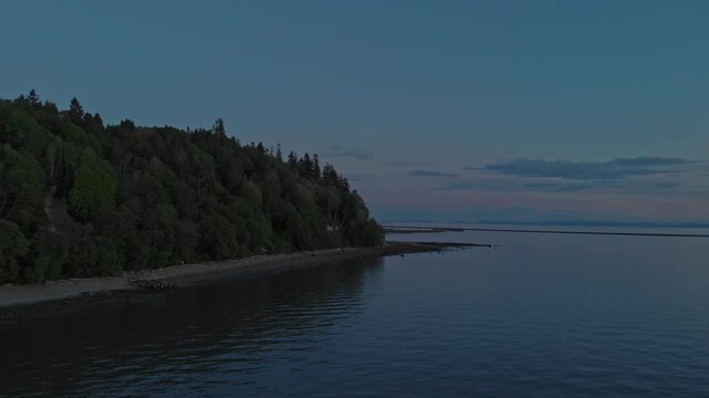 Smooth drone flight glides over calm sea surface tracking around shoreline forest while vibrant pink sky reflects on water during twilight hours at University of British Columbia coastline