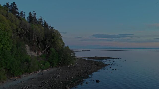 Smooth drone flight reversing over calm sea surface tracking away shoreline forest while vibrant pink sky reflects water during twilight hours at University of British Columbia coastline wreck beach