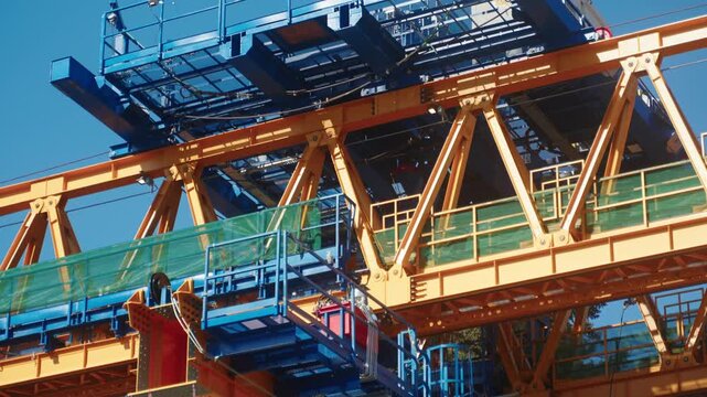 Handheld perspective pans slowly right, providing a detailed close-up view of the complex yellow steel beams, blue supports, and green safety netting of an industrial segment launcher clear blue sky