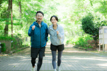 Obraz premium 新緑の公園でウォーキングやランニングのスポーツが楽しいミドル夫婦の正面カメラ目線笑顔 ダイエットや健康などの明るいイメージ全身