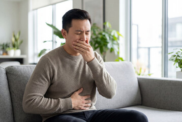 Fototapeta premium Asian man feeling sick with nausea and stomach ache sitting on a sofa at home in the living room