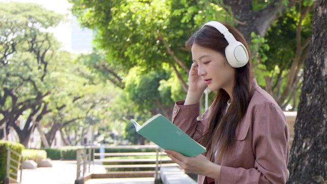 Young Asian Woman Reading Book and Wearing Headphones in Sunny Park