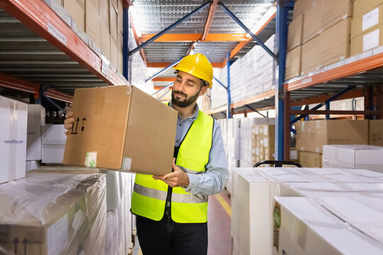 Male warehouse worker wearing safety vest and hardhat, holding cardboard box standing in industrial storage facility. Logistics operations, inventory management, and workflow of distribution center
