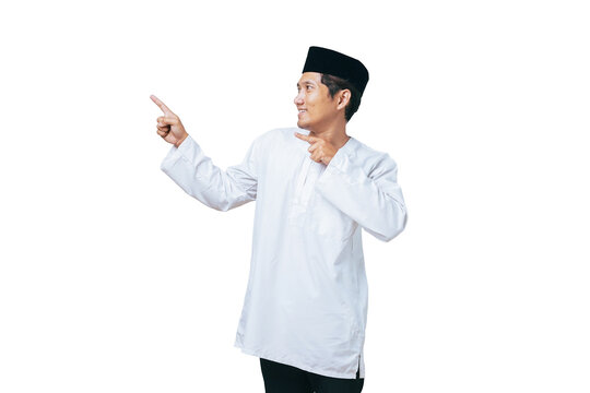 Happy young Muslim man in white baju kurung and black peci pointing both index fingers on his side against white background
