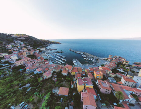 Aerial view of the harbor of Giglio Porto featuring colorful houses, docked boats, and a stone pier along the coastline in Giglio Porto, Tuscany, Italy.