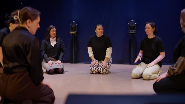 Group of women sitting in a circle on a stage floor practicing interactive theater workshop exercises with expressive body movement and focused communication