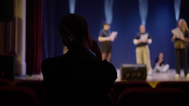 Director observing actors rehearsing on a dimly lit theater stage from the audience seats, evaluating performance during dramatic script reading practice session