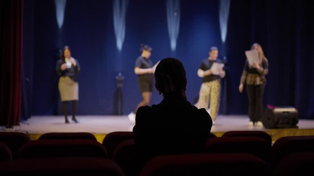 Theatre director observing actors rehearsing lines on a dimly lit stage from the back row of an empty auditorium during an evening performance practice