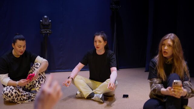 Young adults sitting in a relaxed circle on a stage, chatting and laughing together during an informal rehearsal or creative workshop in a dimly lit studio space