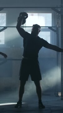 Silhouetted athlete performing powerful kettlebell overhead lift in dimly lit industrial gym with dramatic backlighting and focused strength training intensity