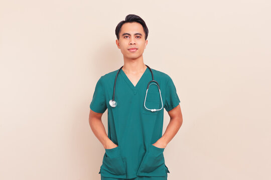 Confident Southeast Asian male doctor in green scrubs with stethoscope and hands in pockets. Professional healthcare worker or medical intern on isolated background.