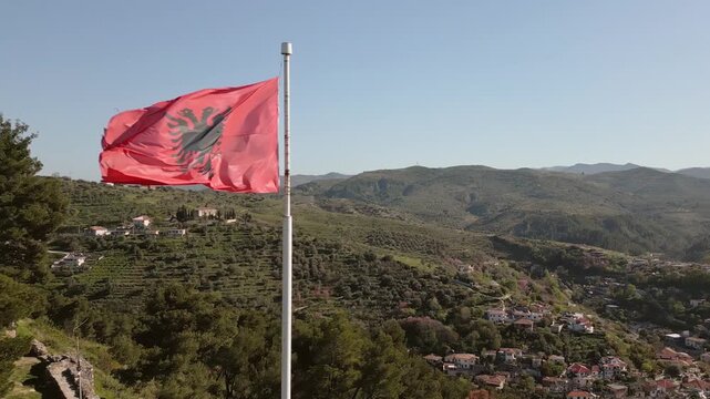 Aerial view Albanian national flag prominently wave atop stone fortification at Berat Castle - Kalaja e Beratit under clear blue sky, overlooking lush, forested hillside and Berat cityscape