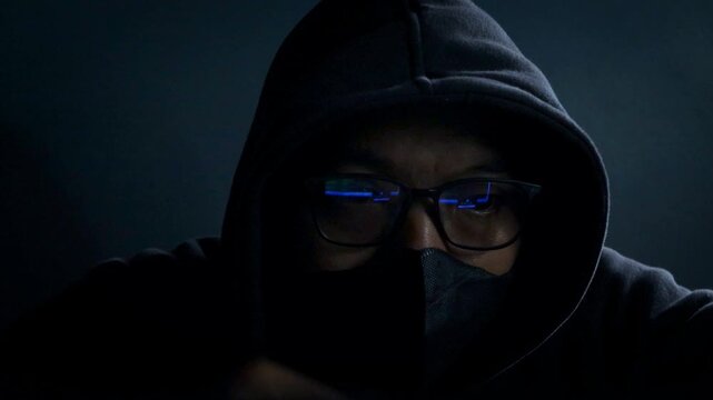 Hacker wearing glasses and face mask conducting cyber attack in a dark room, network breach and ransomware in internet fraud. Night scene with computer, spyware, dark web and online fraud activity.