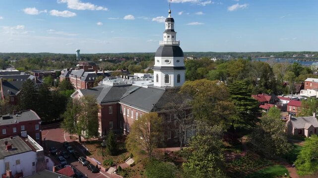 Aerial approaching shot of Annapolis Maryland State House with historic brick buildings and green trees under bright spring sunlight. Calm atmosphere in classic American capital setting. Wide shot