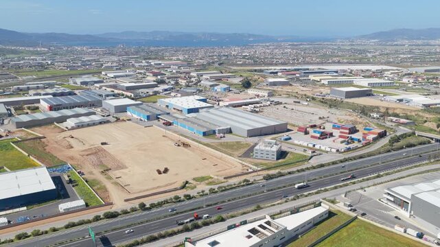 Aerial drone footage circling over a vast industrial area, showcasing warehouses, logistic centers, and a busy highway with traffic, all set against a backdrop of a distant city and sea