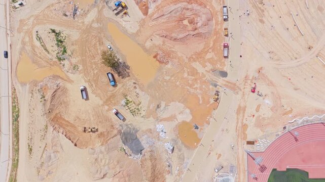 Heavy machinery and dump trucks working on a large construction site with piles of earth and mud, as seen from a top-down aerial perspective next to a running track and road