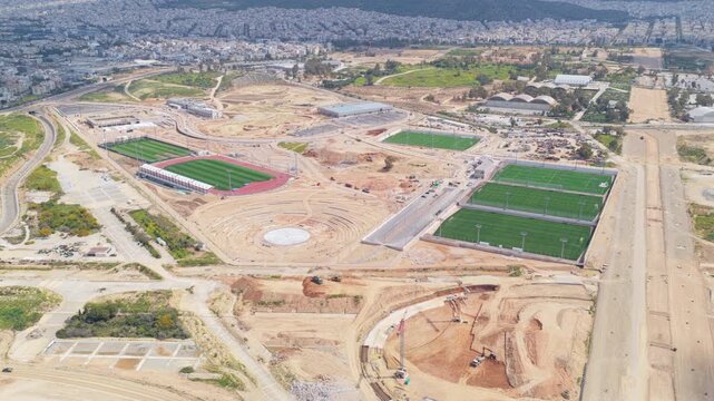 High aerial perspective following the ongoing construction work of a modern sports complex, featuring new football fields, a running track, and an amphitheater surrounded by earthworks