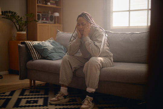 Young adult woman sitting with electronic ankle monitor, holding head on sofa during house arrest. Useful for legal restriction, criminal justice, surveillance, confinement themes
