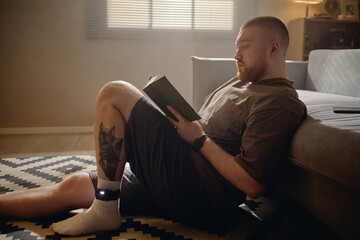 Obraz premium Young man reading book at home while wearing electronic ankle monitor, representing house arrest and criminal justice surveillance. Useful for legal, prison reform, probation, rehabilitation campaigns