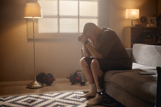 Young adult man sitting with electronic ankle monitor, bowing head and clasping hands at home, representing house arrest and legal restriction. Useful for justice, crime, probation campaigns