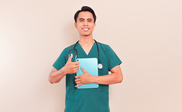 Smiling Southeast Asian male nurse or doctor in green scrubs and stethoscope holding a digital tablet and showing thumbs up. Professional healthcare worker giving approval sign on isolated background.