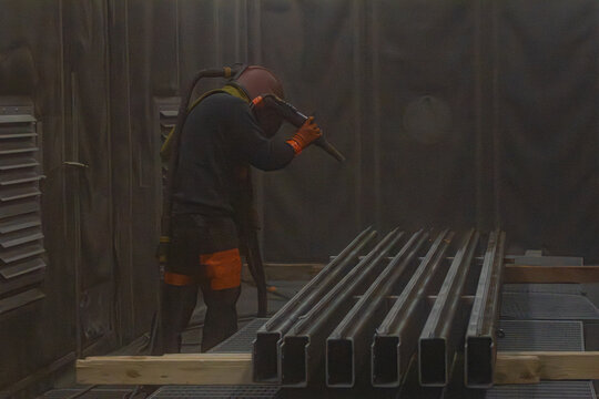 Artisan en combinaison de protection utilisant une lance de sablage &agrave; haute pression pour d&eacute;caper des profil&eacute;s m&eacute;talliques dans une cabine industrielle.