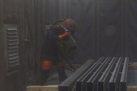 Artisan en combinaison de protection utilisant une lance de sablage &agrave; haute pression pour d&eacute;caper des profil&eacute;s m&eacute;talliques dans une cabine industrielle.