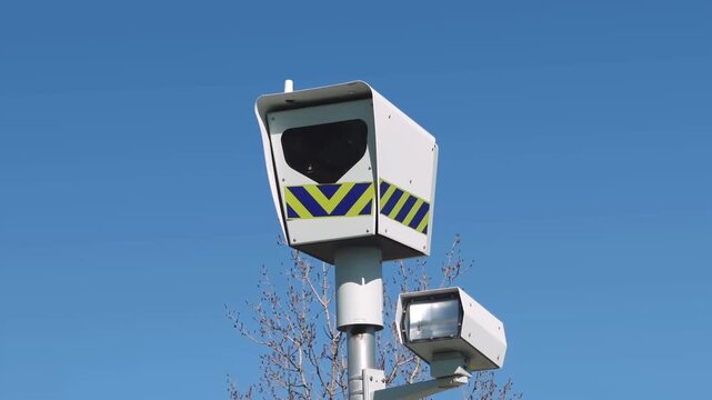 Automated speed enforcement camera and flash unit mounted on a pole against a clear blue sky to monitor traffic and road safety.
