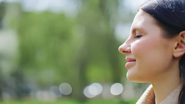 Beautiful woman with a peaceful, happy expression enjoys tranquil moment outdoors. Gentle sunlight illuminates face, conveying contentment and optimism amidst green nature. Thoughtful girl