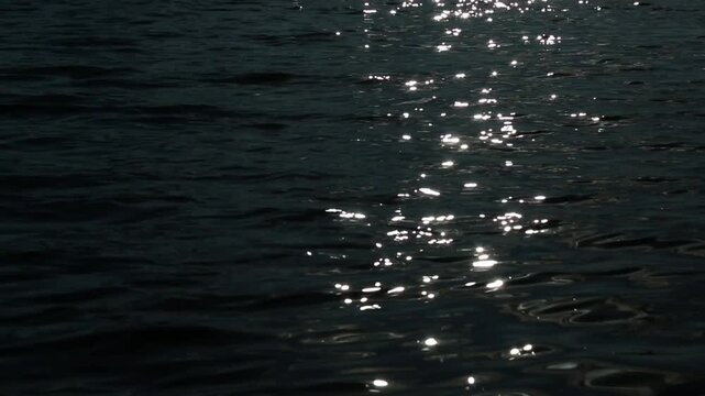 Blue sea water surface filmed in daylight as bright sun path expands and moves across rippling waves from left to right then scene transitions to dark in final frame.