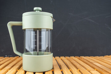 A green French press coffee maker stands on a wooden surface with dark background. The composition emphasizes kitchen lifestyle. © Jakob
