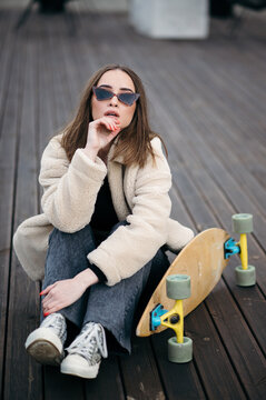 Young woman in stylish outfit with skateboard sitting on wooden deck