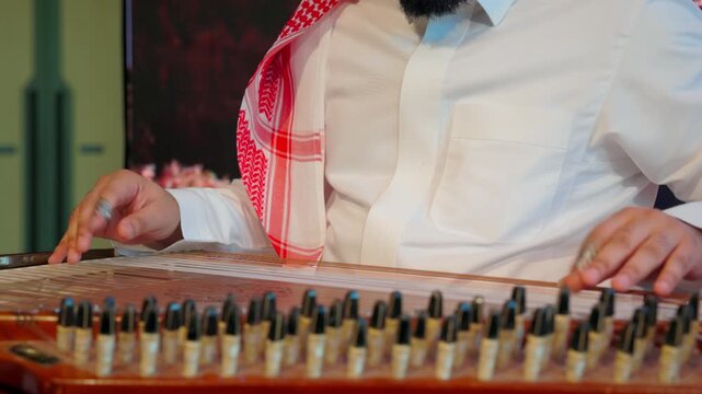 Saudi Arabian musician wearing traditional attire, including a
white thobe and red-and-white ghutra, playing a qanun. The performer&rsquo;s hands
are visible plucking the strings with finger picks.