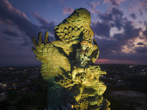 Aerial view of the illuminated Garuda Wisnu Kencana statue under a dramatic sunset sky with city lights in the background Kecamatan Kuta Selatan, Bali, Indonesia.