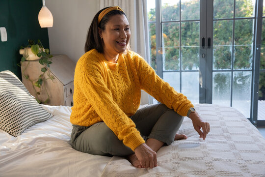 Mature woman sitting cross-legged on bed at home, wearing yellow sweater, touching coverlet