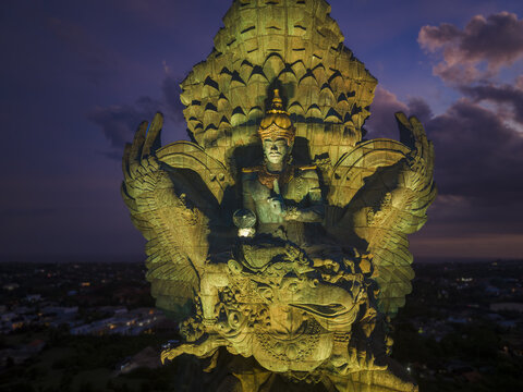 Aerial view of the illuminated Garuda Wisnu Kencana statue at twilight with city lights in the background in Kecamatan Kuta Selatan, Bali, Indonesia.