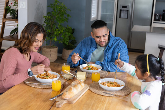 Family of three sitting at wooden dining table sharing pasta with forks and orange juice