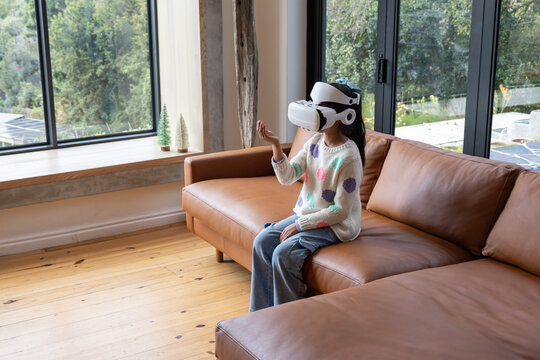 Asian preteen girl sitting on leather sofa at home wearing cream sweater using white VR headset