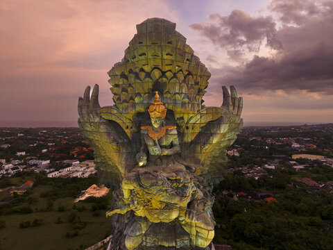 Aerial view of the Garuda Wisnu Kencana statue at sunset with dramatic clouds and the surrounding urban landscape in Kecamatan Kuta Selatan, Bali, Indonesia.