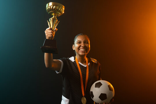 Female athlete holding trophy aloft while wearing soccer jersey and medal, cradling ball in studio