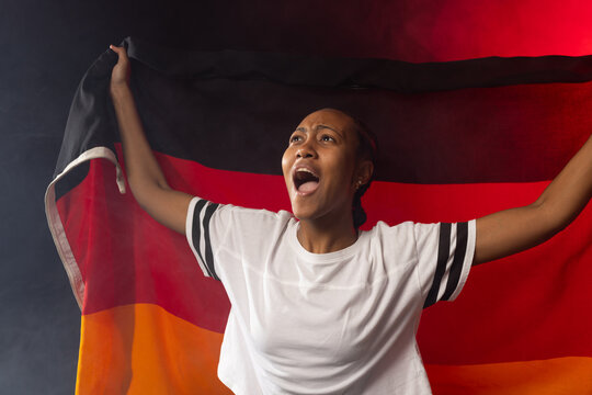African American woman in white tee holding tricolor flag, cheering with red-lit fog in studio