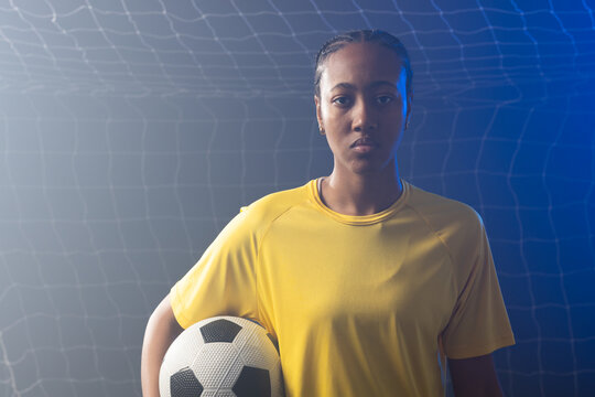 African American female wearing yellow jersey holding soccer ball at right hip in goal net, rim-lit