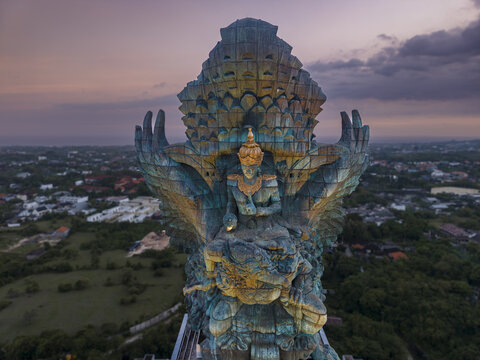 Aerial view of the Garuda Wisnu Kencana statue at sunset with its intricate copper details and the surrounding urban landscape of Kecamatan Kuta Selatan, Bali, Indonesia.