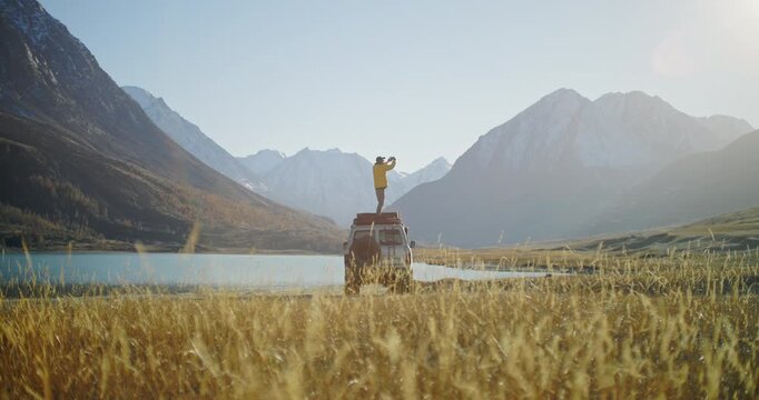 Traveler in a yellow jacket standing on the roof of an off road SUV and taking a smartphone photo near a mountain lake in a remote alpine valley. Snowy peaks, golden grass, and bright daylight create