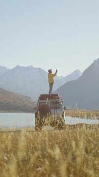 Man in a yellow jacket standing on the roof of an off road vehicle and shooting a phone picture by a mountain lake with snowy peaks in the background. Wide natural landscape with dry grass and bright