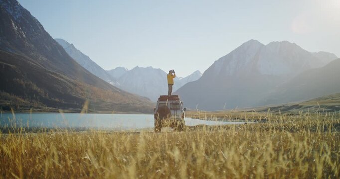 Traveler in a yellow jacket standing on the roof of an off road SUV and taking a smartphone photo near a mountain lake in a remote alpine valley. Snowy peaks, golden grass, and bright daylight create