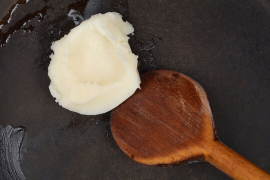 A top down shot of a white dollop of pork lard melting in a dark frying pan with a wooden spoon.