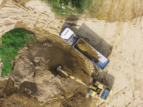 Aerial view of a yellow excavator loading soil into a white dump truck at a construction site with tire tracks and green patches Taiwan.